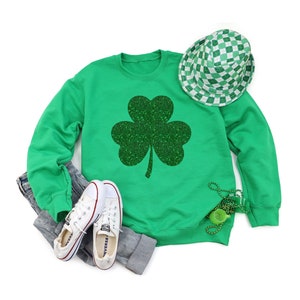 K&ouml;nnte beinhalten: Gr&uuml;nes Sweatshirt mit einem glitzernden gr&uuml;nen Kleeblatt-Design. Das Sweatshirt wird mit wei&szlig;en Turnschuhen und einer gr&uuml;n-wei&szlig; karierten M&uuml;tze kombiniert.