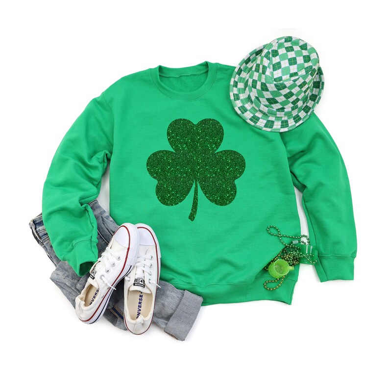 K&ouml;nnte beinhalten: Gr&uuml;nes Sweatshirt mit einem glitzernden gr&uuml;nen Kleeblatt-Design. Das Sweatshirt wird mit wei&szlig;en Turnschuhen und einer gr&uuml;n-wei&szlig; karierten M&uuml;tze kombiniert.