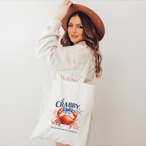 May include: A white tote bag with a crab illustration and the text "if Crabby, Please Return to the Beach." The crab is red and orange with blue accents. The bag has long handles.