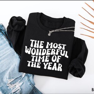 May include: A black long-sleeve sweatshirt featuring the white text THE MOST WONDERFUL TIME OF THE YEAR in a wavy font, paired with light blue distressed denim jeans and a silver chain necklace resting on the neckline of the garment.