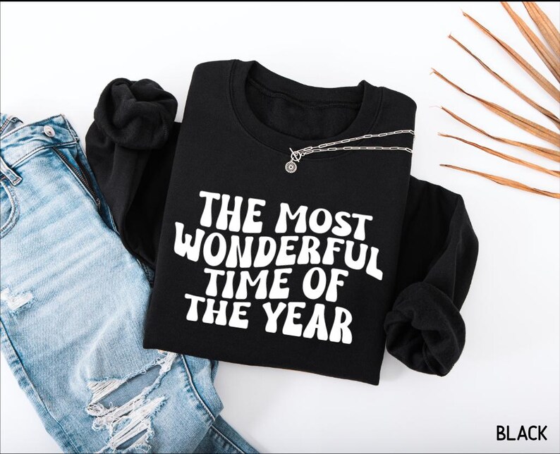 May include: A black long-sleeve sweatshirt featuring the white text THE MOST WONDERFUL TIME OF THE YEAR in a wavy font, paired with light blue distressed denim jeans and a silver chain necklace resting on the neckline of the garment.