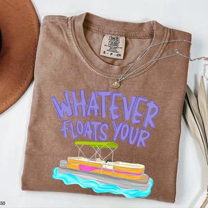 Puede incluir: Camiseta color espresso con la frase "WHATEVER FLOATS YOUR" en letras moradas. Un gr&aacute;fico colorido de un pont&oacute;n est&aacute; debajo del texto. Un collar plateado con un colgante est&aacute; sobre la camiseta.