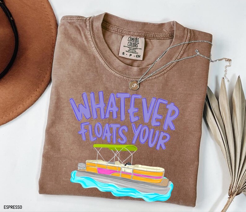Puede incluir: Camiseta color espresso con la frase "WHATEVER FLOATS YOUR" en letras moradas. Un gr&aacute;fico colorido de un pont&oacute;n est&aacute; debajo del texto. Un collar plateado con un colgante est&aacute; sobre la camiseta.
