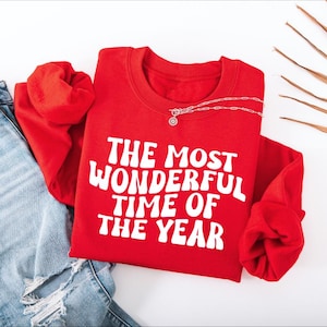 May include: A bright red long-sleeve crewneck sweatshirt featuring the white text THE MOST WONDERFUL TIME OF THE YEAR printed in a bold, wavy font across the chest. The garment is paired with light blue distressed denim jeans.