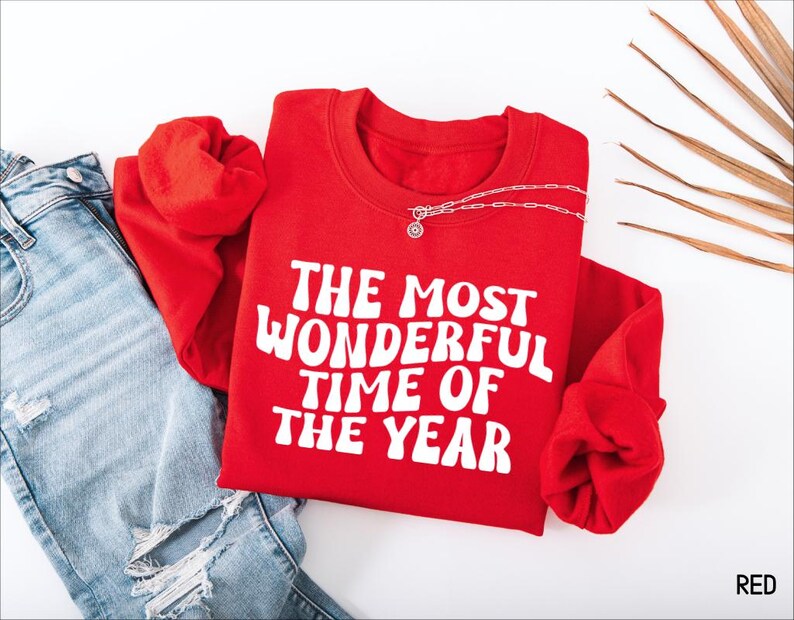 May include: A bright red long-sleeve crewneck sweatshirt featuring the white text THE MOST WONDERFUL TIME OF THE YEAR printed in a bold, wavy font across the chest. The garment is paired with light blue distressed denim jeans.