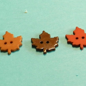 May include: A collection of eight buttons shaped like autumn leaves and one acorn. The buttons are in various shades of red, dark green, orange, brown, and yellow. Each leaf button features two central holes for sewing, while the acorn has one.