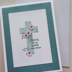 May include: A white card with a teal border and a cross with pink flowers. The text on the card reads "God is, and all is well. -John Whittier".
