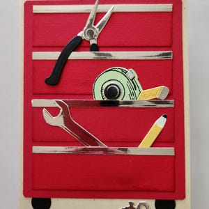 May include: A red toolbox-themed greeting card with silver accents. The card features a pair of pliers, a tape measure, a wrench, and a pencil. The card has a cream-colored border and black wheels.