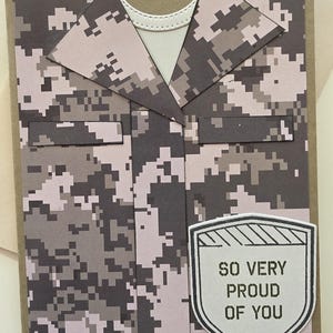 May include: A handmade greeting card featuring a camouflage military uniform design. The card includes a white collar and a shield-shaped emblem with the text "SO VERY PROUD OF YOU" in black. The background is a neutral brown.