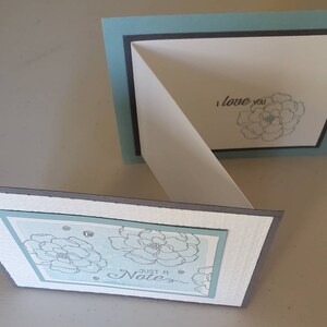 May include: A folded card with a light blue border and a white interior. The front of the card features a floral design with the text "Just a Note". The inside of the card has a single flower design with the text "I love you".