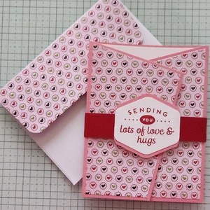 May include: A handmade greeting card with a pink heart pattern and a red band. The card reads "Sending you lots of love & hugs." An envelope with a matching heart pattern is included.
