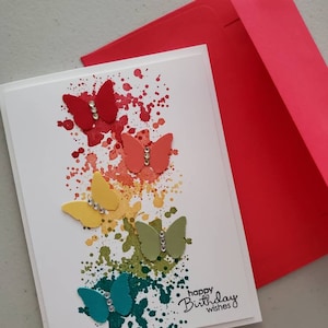 May include: A white card with a rainbow splatter paint background and five colorful paper butterflies with rhinestones. The card says "happy birthday wishes".
