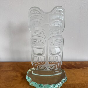 May include: A clear glass totem pole sculpture with etched Native American-inspired designs. The sculpture has a rectangular shape with a rounded top and is mounted on a clear, irregular-shaped base. The artwork is displayed on a wooden surface.