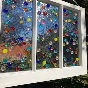 May include: A white painted window frame with three panes of glass. Each pane is decorated with colourful glass marbles of various sizes and shapes.