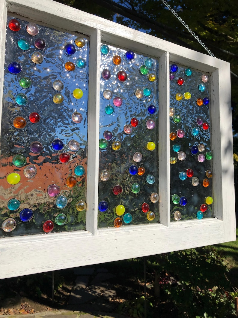 May include: A white painted window frame with three panes of glass. Each pane is decorated with colourful glass marbles of various sizes and shapes.