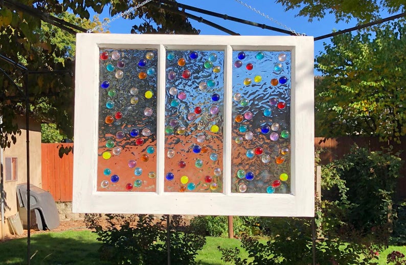 May include: A white window frame with three panes of glass, each filled with colourful glass beads. The beads are arranged in a random pattern and create a whimsical and colourful design.