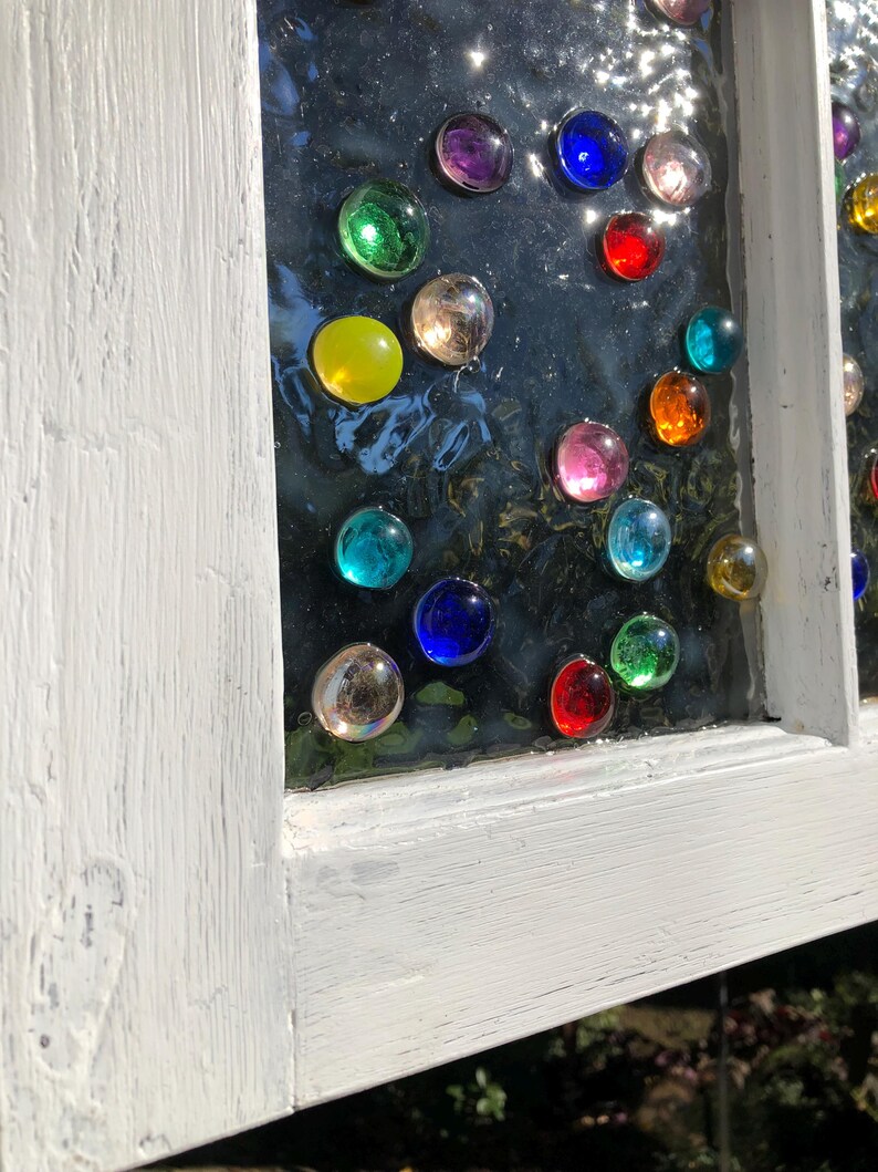 May include: A white painted window frame with a glass pane featuring a colourful mosaic of round glass gems. The gems are arranged in a circular pattern and include shades of red, blue, green, yellow, and purple.