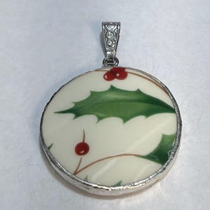 May include: A round pendant featuring a cream-colored background with a painted green holly leaf and red berry design. The circular piece is encased in a silver-toned metal border and includes a decorative silver-toned metal bail at the top.