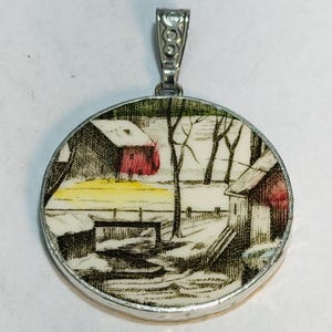 May include: A circular pendant featuring a detailed black and white ink-style illustration of a winter landscape with red buildings, a yellow horizon line, and bare trees, encased in a silver-toned metal frame with a decorative bail at the top.