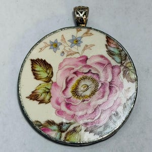 May include: A circular pendant featuring a cream-colored background with a large, detailed pink rose illustration at the center. The design includes small blue flowers and green leaves, all encased in a thin silver-toned metal frame with a decorative bail.