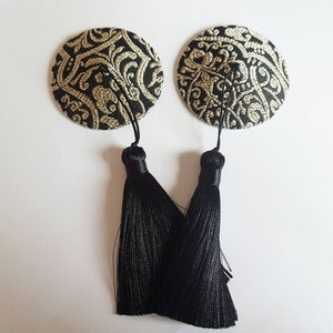 May include: Two round, black and gold fabric nipple pasties with a detailed, ornate pattern. Each pastie has a black tassel hanging from the bottom. The background is white.