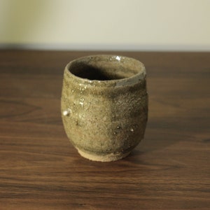 May include: A small, rustic, brown ceramic cup with a textured surface. The cup is slightly wider at the top than the bottom.