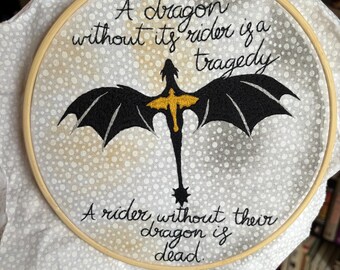 Puede incluir: Arte de bordado en aro con una silueta negra de un dragón con detalles amarillos. El texto dice "Un dragón sin su jinete es una tragedia. Un jinete sin su dragón está muerto."