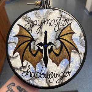 May include: Embroidered hoop art featuring the words "Spymaster Shadowsinger" in black script. The design includes a black sword with blue accents and gold and black dragon wings on a light blue and white patterned background.