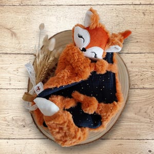 May include: A plush orange fox-shaped baby blanket with a navy blue interior dotted with gold stars. The fox has a white face, ears, and tail tip. The blanket is displayed on a wooden slice with decorative dried flowers and ribbon.