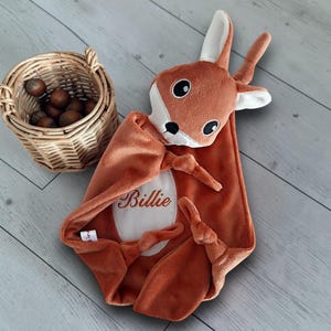 Peut inclure: Une couverture de sécurité en peluche douce représentant une tête de cerf couleur rouille avec des accents blancs et des yeux noirs brodés. Le corps de la couverture est rouille avec un ovale blanc central brodé du nom Billie en brun.