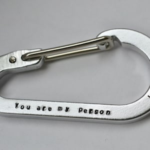 以下が含まれることがあります： 「You are my Person」の刻印と「K <3 J」のイニシャルが入ったシルバーのカラビナクリップ。クリップにはスプリング式のゲートがあり、アイテムを取り付けるように設計されています。