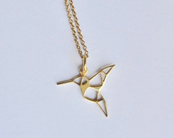May include: A gold-colored hummingbird pendant necklace. The pendant is a geometric design, with a detailed outline of a hummingbird. The necklace chain is delicate and gold-toned. The background is white, with a brown branch in the foreground.