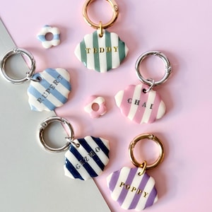 Puede incluir: Una colección de etiquetas para mascotas con nombres como "TEDDY", "RUPERT", "CHAI", "GIZMO" y "POPPY". Cada etiqueta tiene una forma única, rayas y un cierre de metal. Las etiquetas son de varios colores, incluyendo azul, rosa y morado, sobre un fondo rosa.