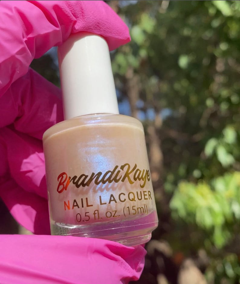 Peut inclure: Un flacon de vernis &agrave; ongles BrandiKaye. Le flacon transparent contient un vernis beige nacr&eacute;. L'&eacute;tiquette dor&eacute;e indique "BrandiKaye NAIL LACQUER" et le volume, 15 ml. Le flacon a un bouchon blanc.