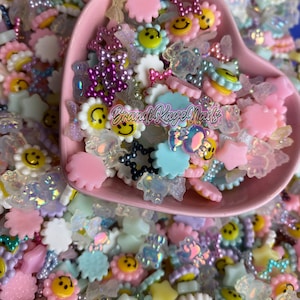 May include: A pink heart-shaped bowl filled with colorful resin charms. The charms include smiley faces, stars, flowers, and butterflies. The charms are perfect for adding a touch of whimsy to your crafts.