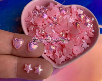 May include: Pink iridescent heart and star shaped nail charms in a pink heart shaped bowl.  The charms are scattered on a table.