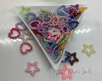 May include: A variety of iridescent pearl-like beads in the shapes of hearts and stars. The beads are arranged in a white triangular dish. The text "BrandiKayeNails" is visible in the bottom right corner of the image.