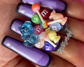 May include: Purple long acrylic nails with 3D candy decorations, including gummy bears, lollipops, and M&Ms. The nails are decorated with black outlines and white lines.