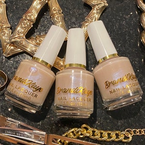 Peut inclure: Trois flacons de vernis &agrave; ongles Brandi Kaye de teinte neutre, chacun avec un bouchon blanc et des lettres dor&eacute;es. Les flacons sont entour&eacute;s de bijoux dor&eacute;s, dont des cr&eacute;oles et une cha&icirc;ne, sur une surface sombre et textur&eacute;e.