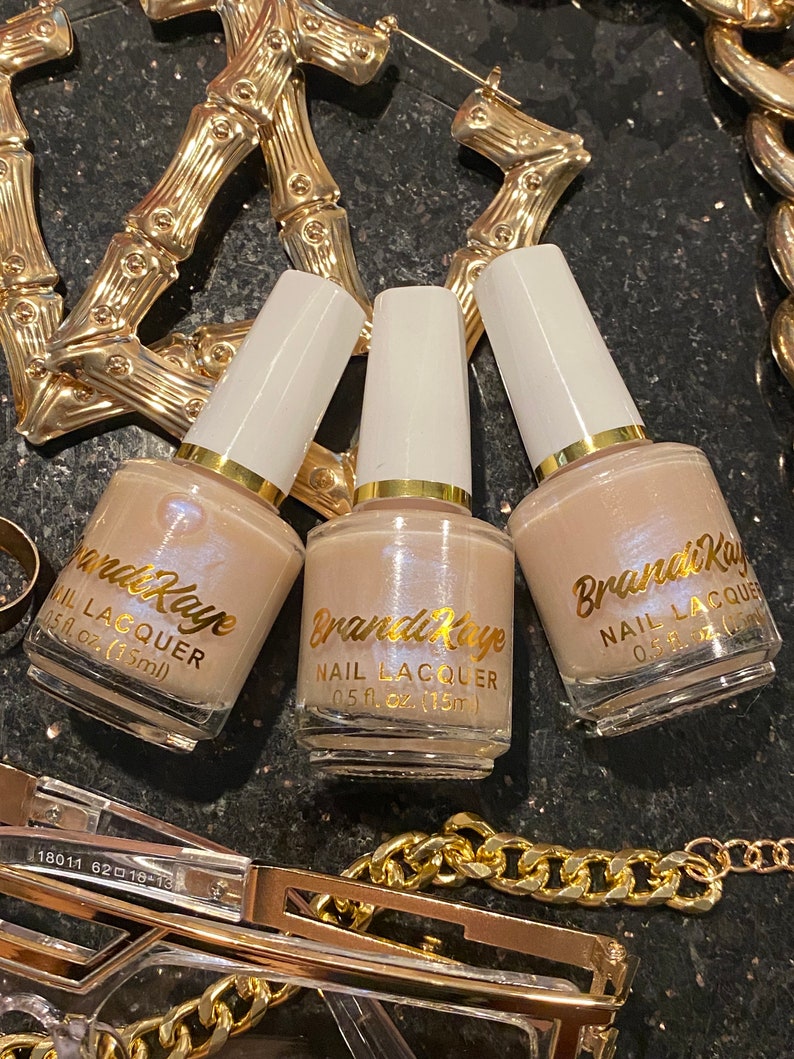 Peut inclure: Trois flacons de vernis &agrave; ongles Brandi Kaye de teinte neutre, chacun avec un bouchon blanc et des lettres dor&eacute;es. Les flacons sont entour&eacute;s de bijoux dor&eacute;s, dont des cr&eacute;oles et une cha&icirc;ne, sur une surface sombre et textur&eacute;e.