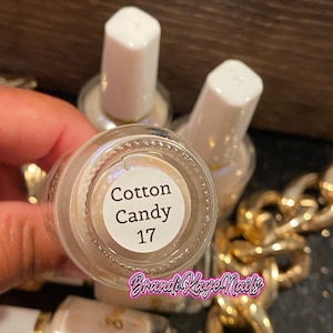 Peut inclure: Gros plan sur plusieurs flacons de vernis &agrave; ongles. Le fond d'un flacon est visible, affichant le texte "Cotton Candy 17". Les flacons ont des bouchons blancs et une couleur de vernis beige clair. La marque "Brandi Kaye Nails" est &eacute;galement visible.