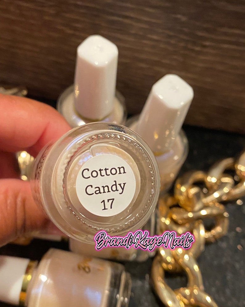 Peut inclure: Gros plan sur plusieurs flacons de vernis &agrave; ongles. Le fond d'un flacon est visible, affichant le texte "Cotton Candy 17". Les flacons ont des bouchons blancs et une couleur de vernis beige clair. La marque "Brandi Kaye Nails" est &eacute;galement visible.