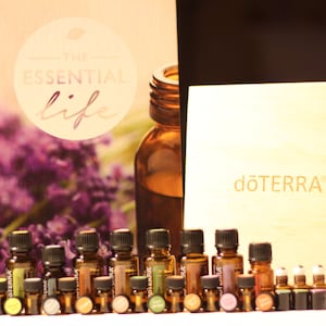 May include: A collection of essential oil bottles in various sizes and colors, arranged in front of a book titled "The Essential Life" and a wooden box with the "dōTERRA" logo. The bottles are amber-colored, with different colored labels.