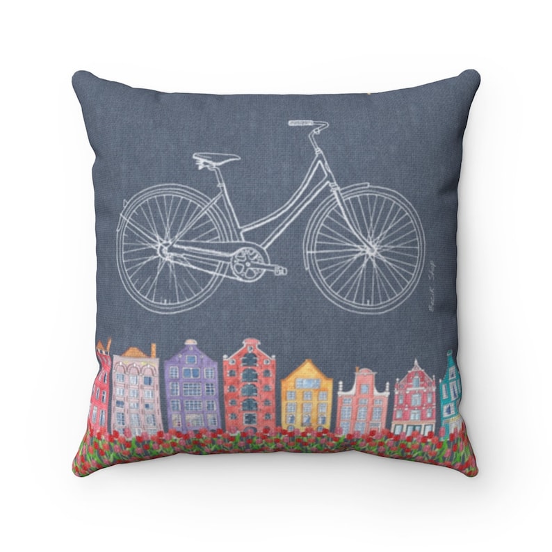 May include: A decorative throw pillow with a blue background featuring a white outline of a bicycle and a row of colorful houses with red flowers at the bottom.