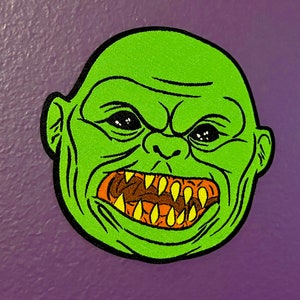 Peut inclure: Visage de monstre vert avec une bouche grande ouverte montrant des dents pointues. Le monstre a un visage vert avec des yeux noirs et un contour noir.