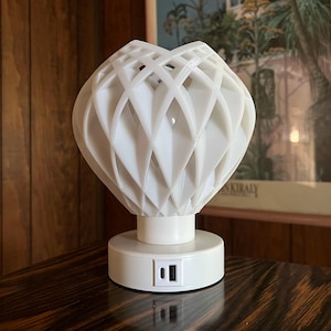 May include: White geometric lamp with a USB charging port on the base. The lamp is on a dark wood surface.