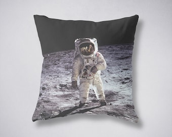 May include: A black and white photograph of an astronaut standing on the moon. The astronaut is wearing a white spacesuit with a helmet and a US flag patch on the left shoulder. The surface of the moon is visible in the background.