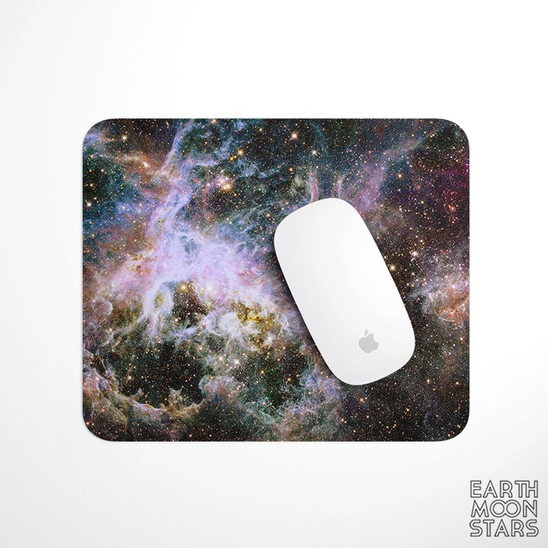 Puede incluir: Una alfombrilla de rat&oacute;n rectangular con un dise&ntilde;o de nebulosa vibrante en tonos morados, azules y dorados. Un rat&oacute;n inal&aacute;mbrico blanco con el logotipo de Apple descansa sobre ella. Accesorio de ordenador.
