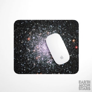 May include: A rectangular mousepad featuring a vibrant image of a galaxy with numerous stars in shades of white, pink, and orange against a black background. A white computer mouse with the Apple logo rests on the pad. The words "EARTH MOON STARS" are printed in white.
