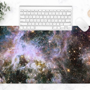 Puede incluir: Alfombrilla de rat&oacute;n rectangular con una imagen vibrante de una nebulosa con tonos morados, azules y dorados. Un teclado y un rat&oacute;n blancos est&aacute;n colocados encima de la alfombrilla. Tambi&eacute;n se ven una taza de caf&eacute; y una planta.